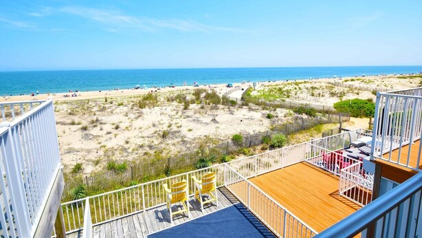 Vista a la playa desde un balcón con terraza de madera, mobiliario exterior y vistas al mar y la costa.