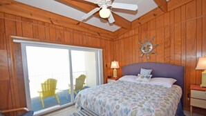 Habitación con paneles de madera, una cama, ventilador de techo, mesita de noche, lámpara y balcón con sillas.
