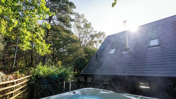 Un bain à remous situé dans une zone boisée, avec une terrasse en bois et un bâtiment doté d’un toit en pente.