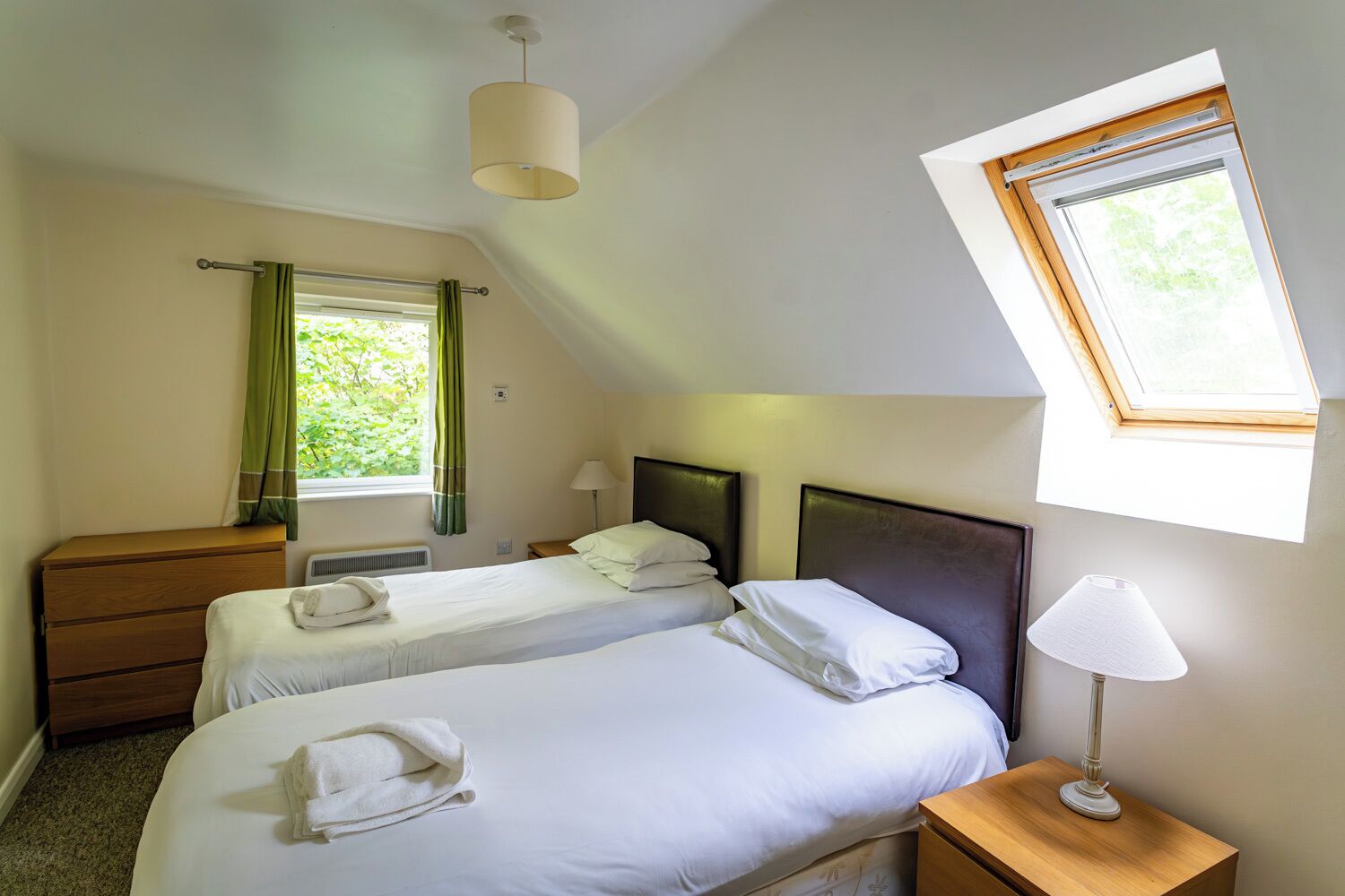 Une chambre avec deux lits, une commode en bois, une table de chevet avec une lampe, une fenêtre donnant sur un paysage verdoyant et un luminaire au plafond.