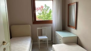 A small, clean room with two single beds, a white radiator, a small table, and a window with a view of trees.