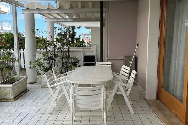 Un patio techado con una mesa y sillas blancas, rodeado de macetas con plantas y vistas a la ciudad.