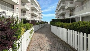 A pathway flanked by white fences and greenery, leading to a row of multi-story residential buildings with balconies.