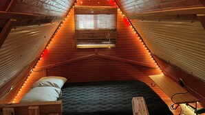 A cozy attic bedroom with a sloped ceiling, wooden walls, a bed with a black mattress, and a small window with blinds. - Beautiful Cabin in a great neighborhood and minutes from Baton Rouge!! (Denham Springs)