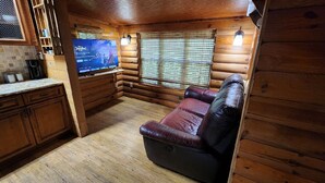 A log cabin interior with a wooden floor, a leather sofa, a television, and a window with blinds. - Beautiful Cabin in a great neighborhood and minutes from Baton Rouge!! (Denham Springs)