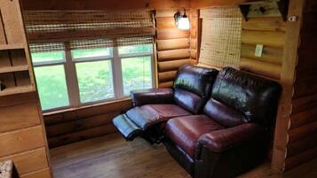 Een interieur van een blokhut met een leren bank, houten planken en een raam met uitzicht op een grasveld.