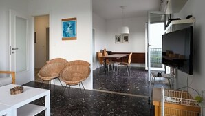 A modern living room with a dining area, a TV, and a balcony with a railing. - VILLA MARGHERITA 1 (Lido di Jesolo)