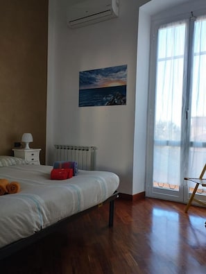 A bedroom with a bed, a radiator, a painting of a sunset, a window with curtains, and a wooden floor. - Sea And Park-genoa Nervi - (Genoa)