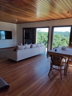 Una sala de estar moderna con un sofá blanco, suelo de madera y ventanas amplias con vistas a un paisaje boscoso.