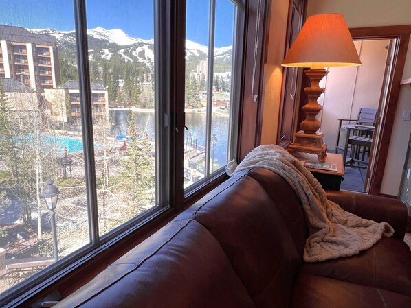 A room with a large window offering a view of a mountain, a pool, and a building.