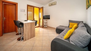 Un soggiorno con un divano grigio, cuscini gialli, un tavolino in legno, una televisione e una porta scorrevole che dà su un balcone.