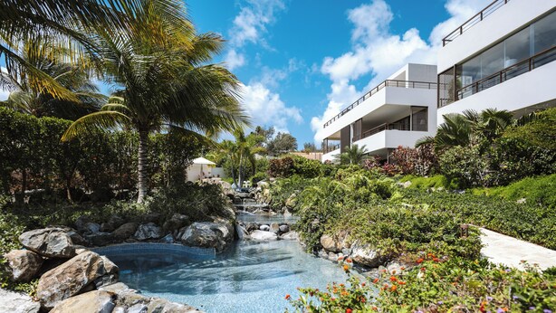 Un complexe hôtelier doté d’une piscine entourée de rochers et de plantes tropicales, d’un bâtiment moderne avec des balcons, et d’un ciel bleu azur.