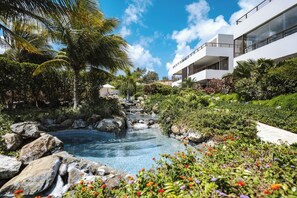 Un complexe hôtelier doté d’une piscine entourée de rochers et de plantes tropicales, d’un bâtiment moderne avec des balcons, et d’un ciel bleu azur.