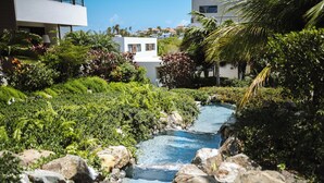 Un jardin paisible avec un petit ruisseau, une végétation luxuriante et un bâtiment moderne en arrière-plan.