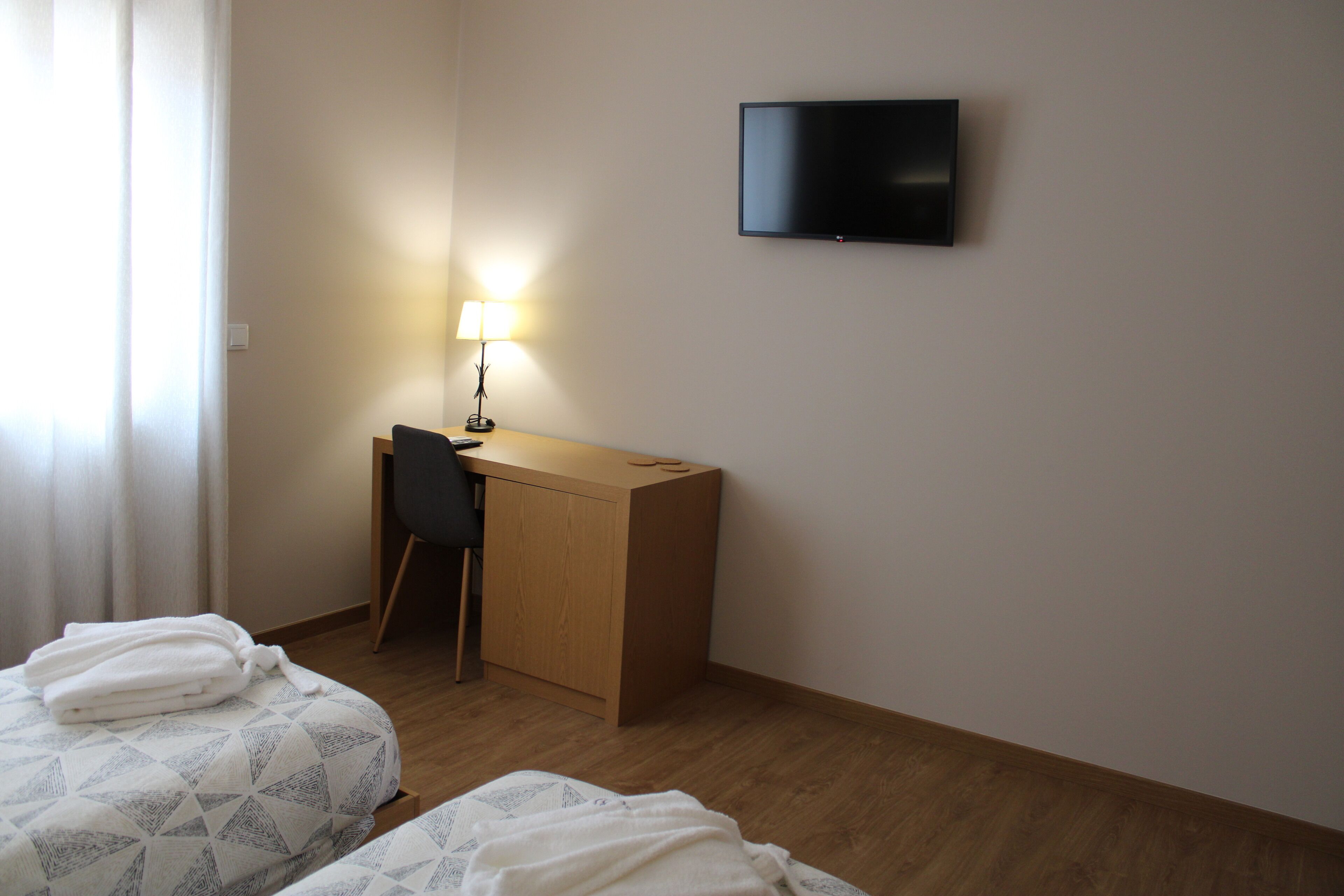 Ein Hotelzimmer mit zwei Betten, einem Schreibtisch mit Stuhl, einem an der Wand montierten Fernseher und einer Schreibtischlampe.