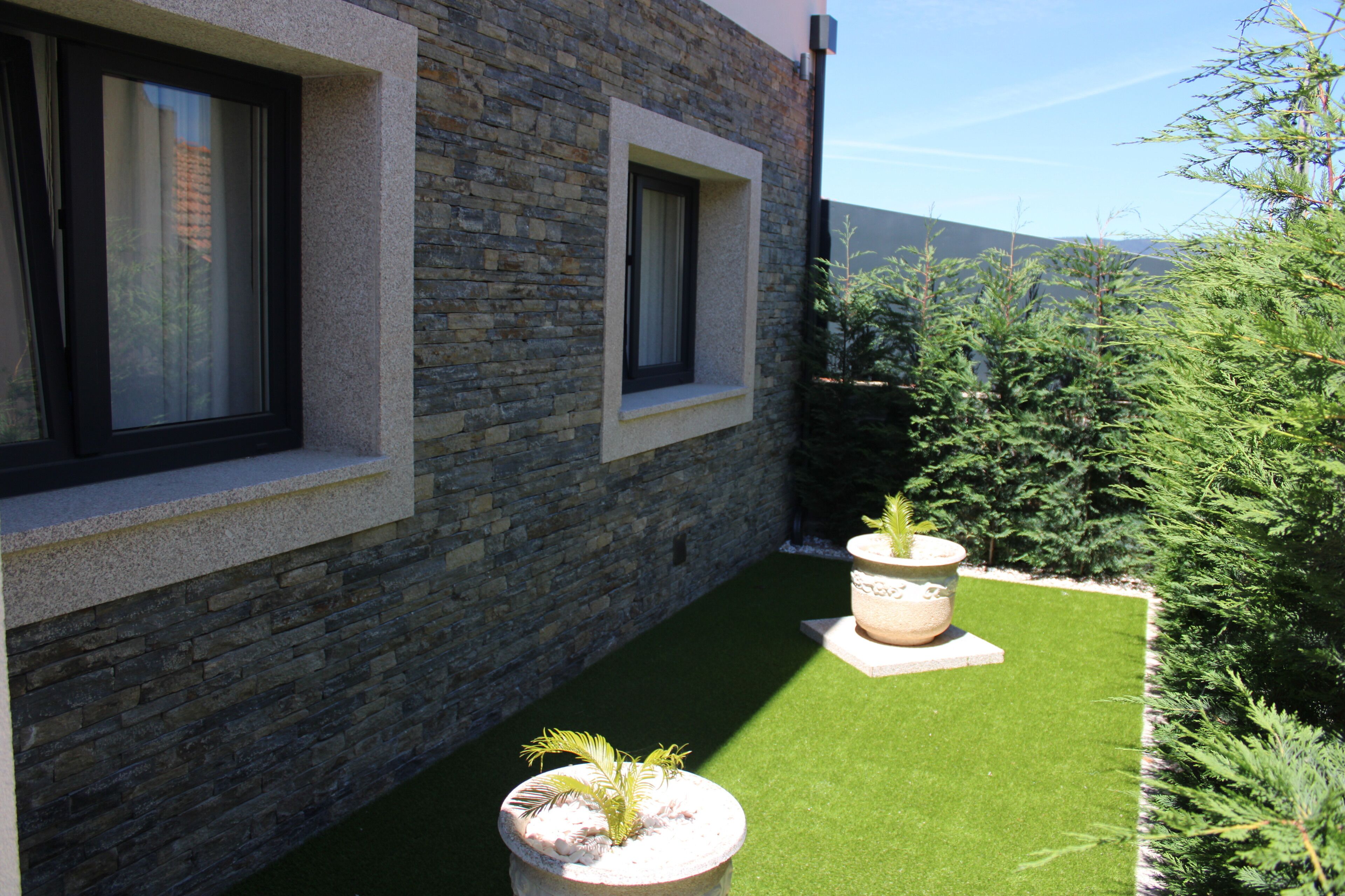 Ein modernes Gebäude mit Steinwänden und großen Fenstern. Vor der Fassade befinden sich zwei Pflanzkästen mit Pflanzen auf einer grünen Rasenfläche.