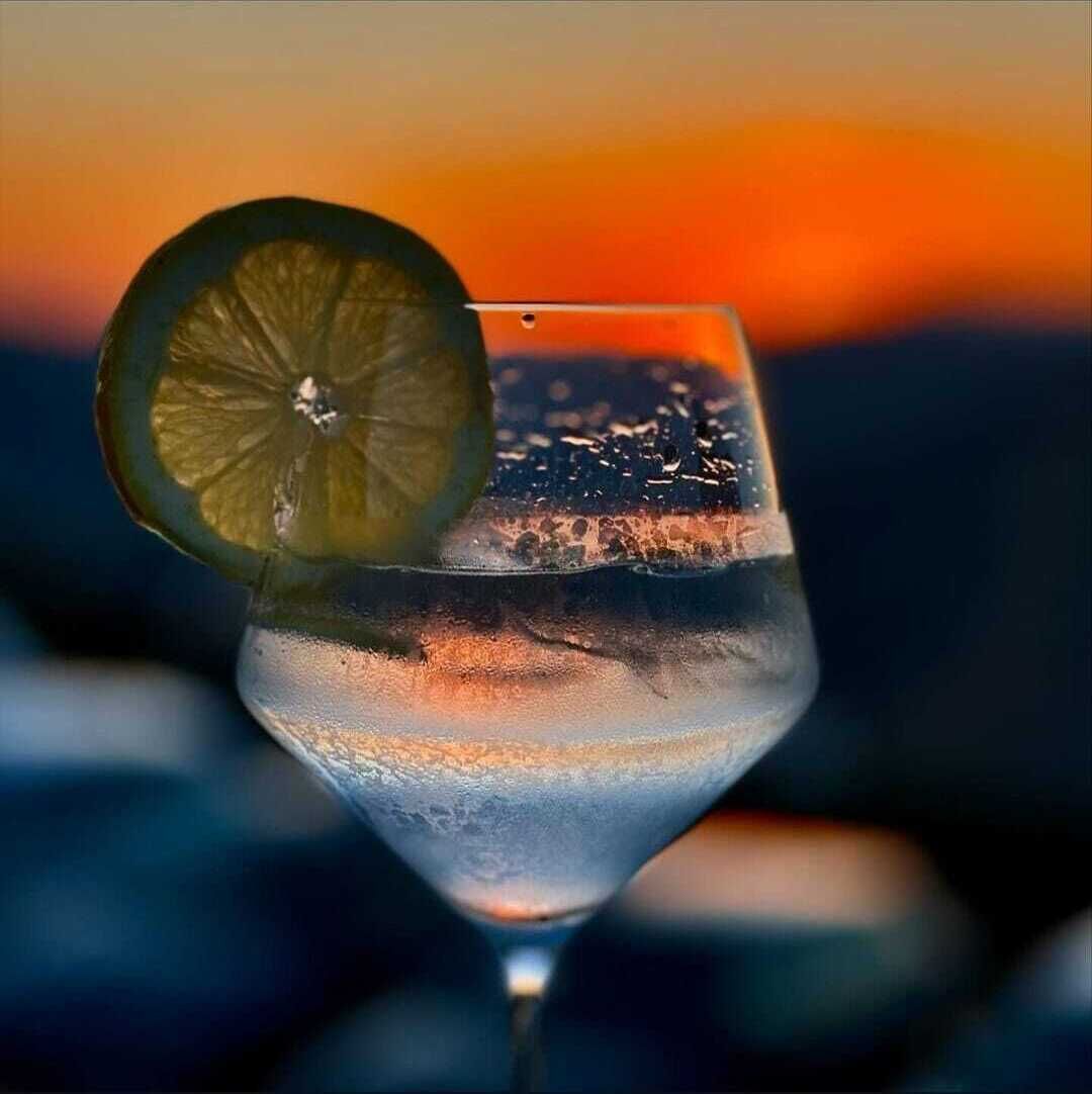 Een cocktailglas met een stukje limoen en ijsblokjes, voor een achtergrond van een zonsondergang.