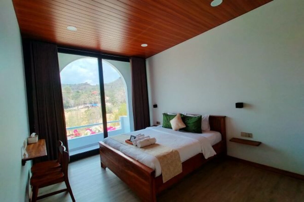 A bedroom with a wooden ceiling, a large window with curtains, a bed with pillows, a bedside table, and a chair.
