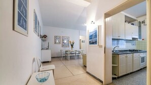 A compact kitchen with white cabinets, a dining area with a table and chairs, and a small seating area with a chair and a basket. - VILLA TERESA 19 (Lido di Jesolo)