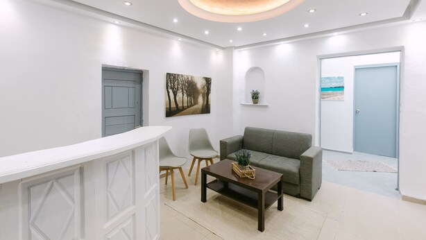 A reception area with a white reception desk, grey chairs, a dark wooden coffee table, a grey sofa, and a painting on the wall. - Seven Grapes - Karterados (Santorini)