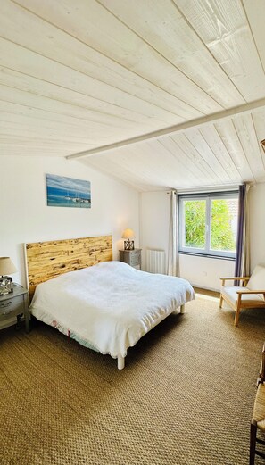 A bedroom with a wooden headboard, a bed with a white blanket, a nightstand with a lamp, a chair, and a window with curtains.