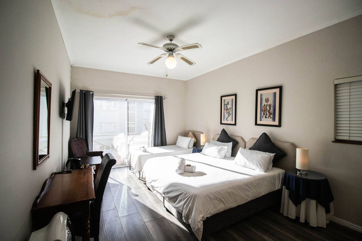 A hotel room with two beds, a desk, a chair, a ceiling fan, and a window with curtains.