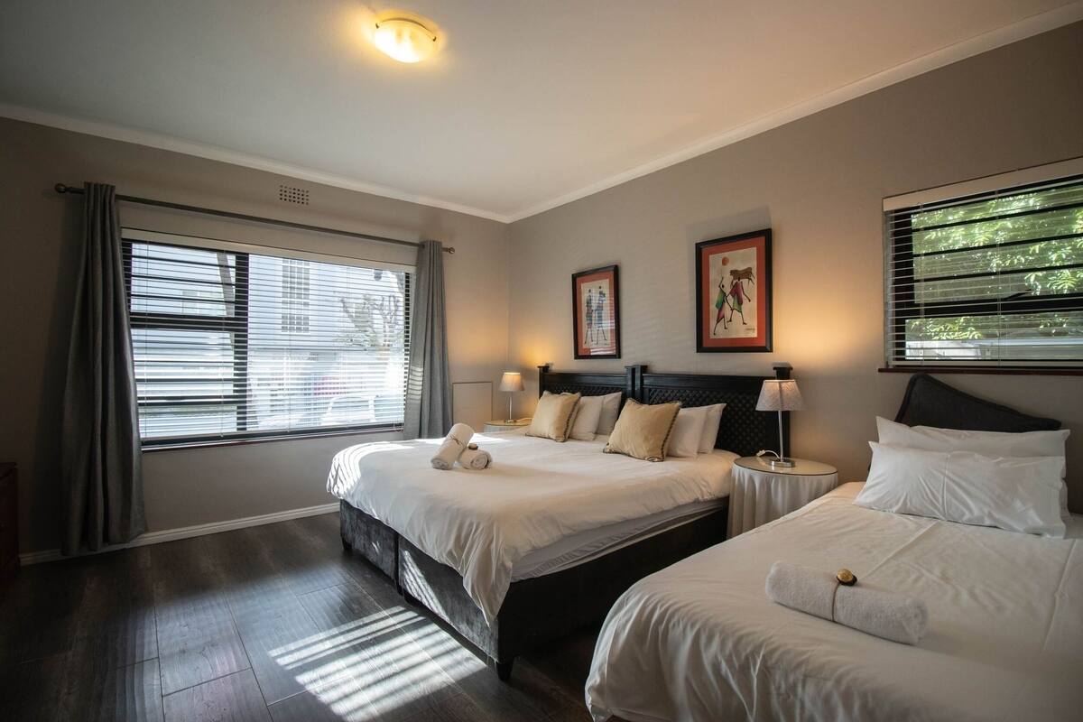 A hotel room with two beds, a large window with blinds, a ceiling light, and two framed pictures on the wall.