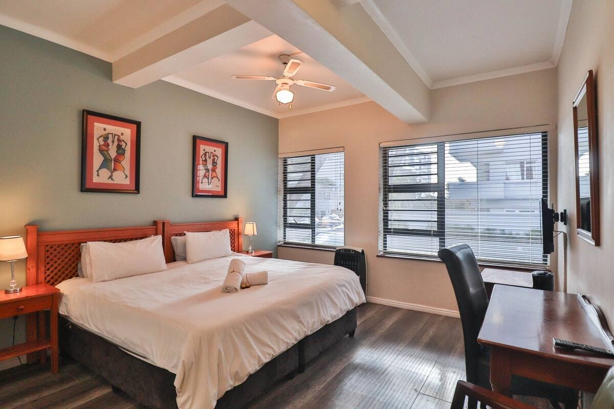 A hotel room with a large bed, a desk, a chair, a ceiling fan, and two windows with blinds.