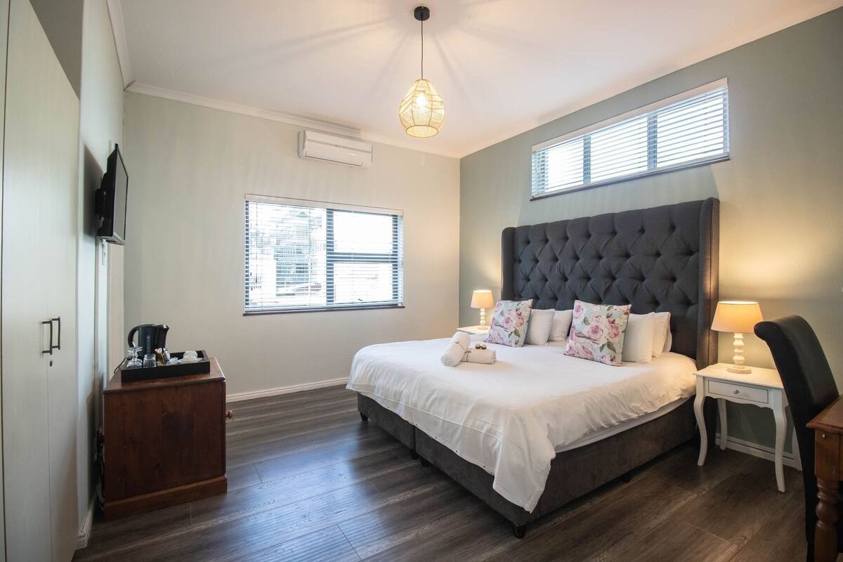 A bedroom with a large bed, a nightstand, a dresser, a TV mounted on the wall, and two windows with blinds.