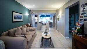 Un salón espacioso con un sofá color beige, una mesa de centro y un cuadro en la pared. Se aprecia una cocina al fondo con una mesa y sillas de comedor.