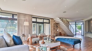 Un salon spacieux avec un grand canapé, un pouf bleu et une table basse en verre. Un escalier mène à l’étage supérieur, et un tapis à motifs zébrés recouvre le sol.