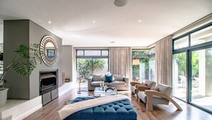 Un salon spacieux avec un grand pouf bleu, un canapé gris et un fauteuil en bois aux lignes courbes. On y trouve une cheminée surmontée d’un miroir moderne, ainsi qu’une grande fenêtre offrant une vue sur l’extérieur.