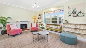 Un salon avec une cheminée, deux fauteuils roses, une table basse ronde et un pouf bleu. Une grande fenêtre offre une vue sur la verdure à l’extérieur.