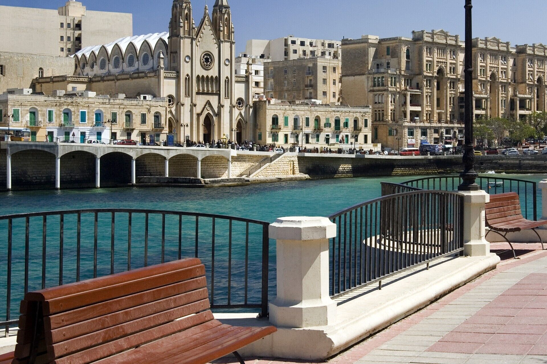 A waterfront promenade with benches and a bridge leading to a historic building with a clock tower.