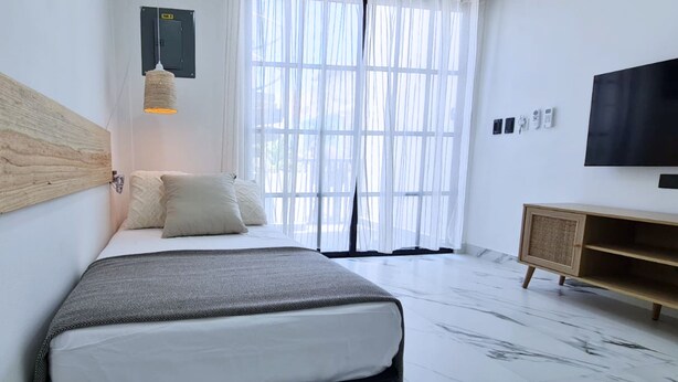 Une chambre à coucher moderne avec un lit, une tête de lit en bois, un téléviseur fixé au mur et une fenêtre ornée de rideaux légers.