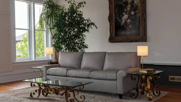 A living room with a grey sofa, a glass coffee table, two lamps, a large plant, and a framed painting on the wall.