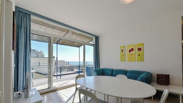 Een kamer met een witte tafel en stoelen, een blauwe bank en een balkon met uitzicht op zee.