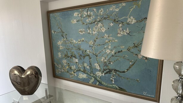 A room with a glass table displaying magazines, a heart-shaped object, and a framed painting of a tree with white blossoms.