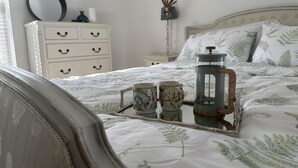 A neatly made bed with a fern leaf pattern, a tray with two mugs, a vintage-style coffee press, and a white dresser with decorative handles.