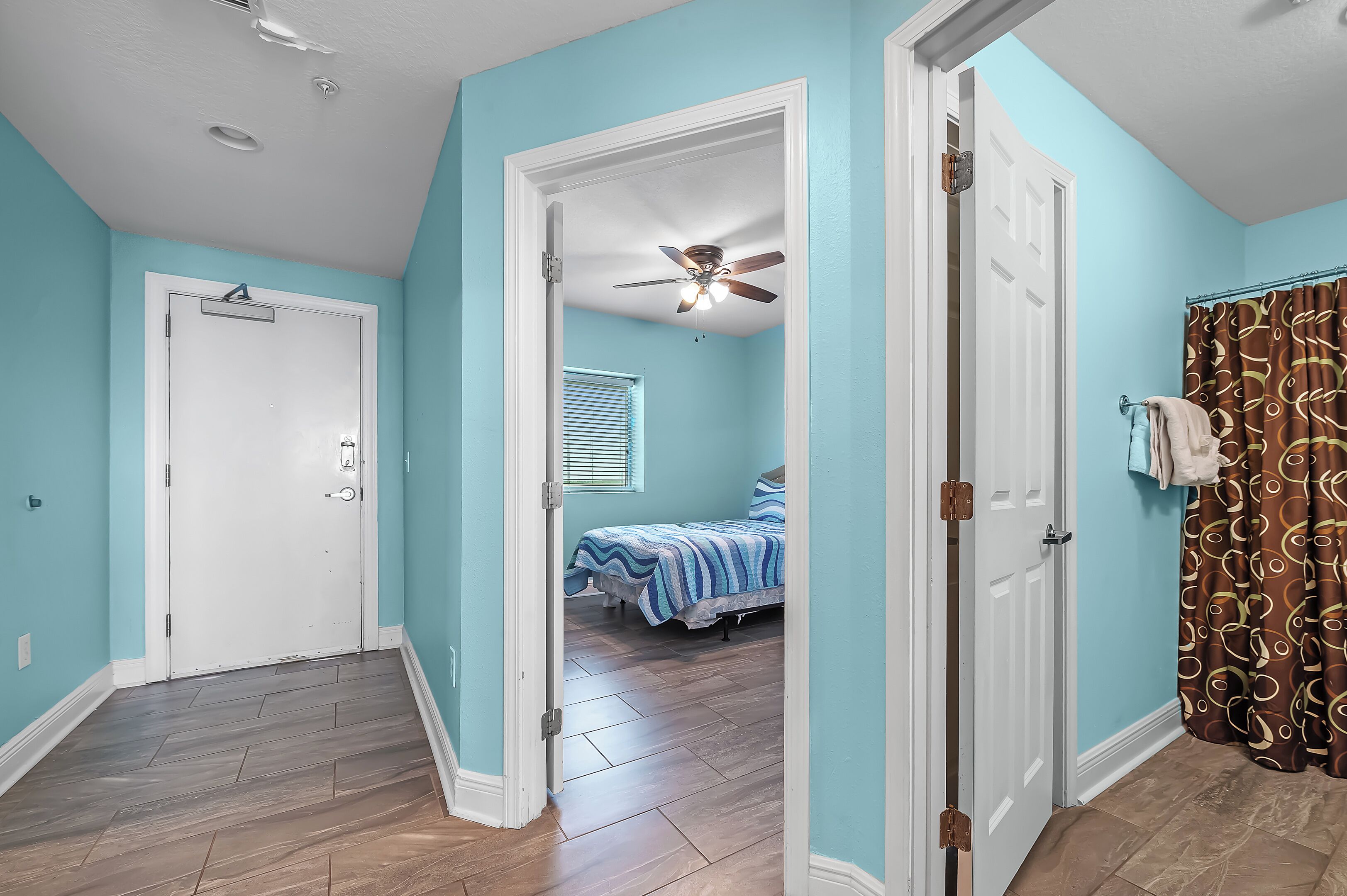 A room with teal walls, a wooden floor, a bed with striped bedding, a ceiling fan, and a shower with a patterned curtain.