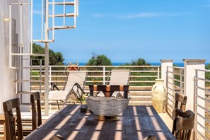 A wooden table with a decorative bowl in the center, surrounded by chairs, set on a patio with a view of the sea.