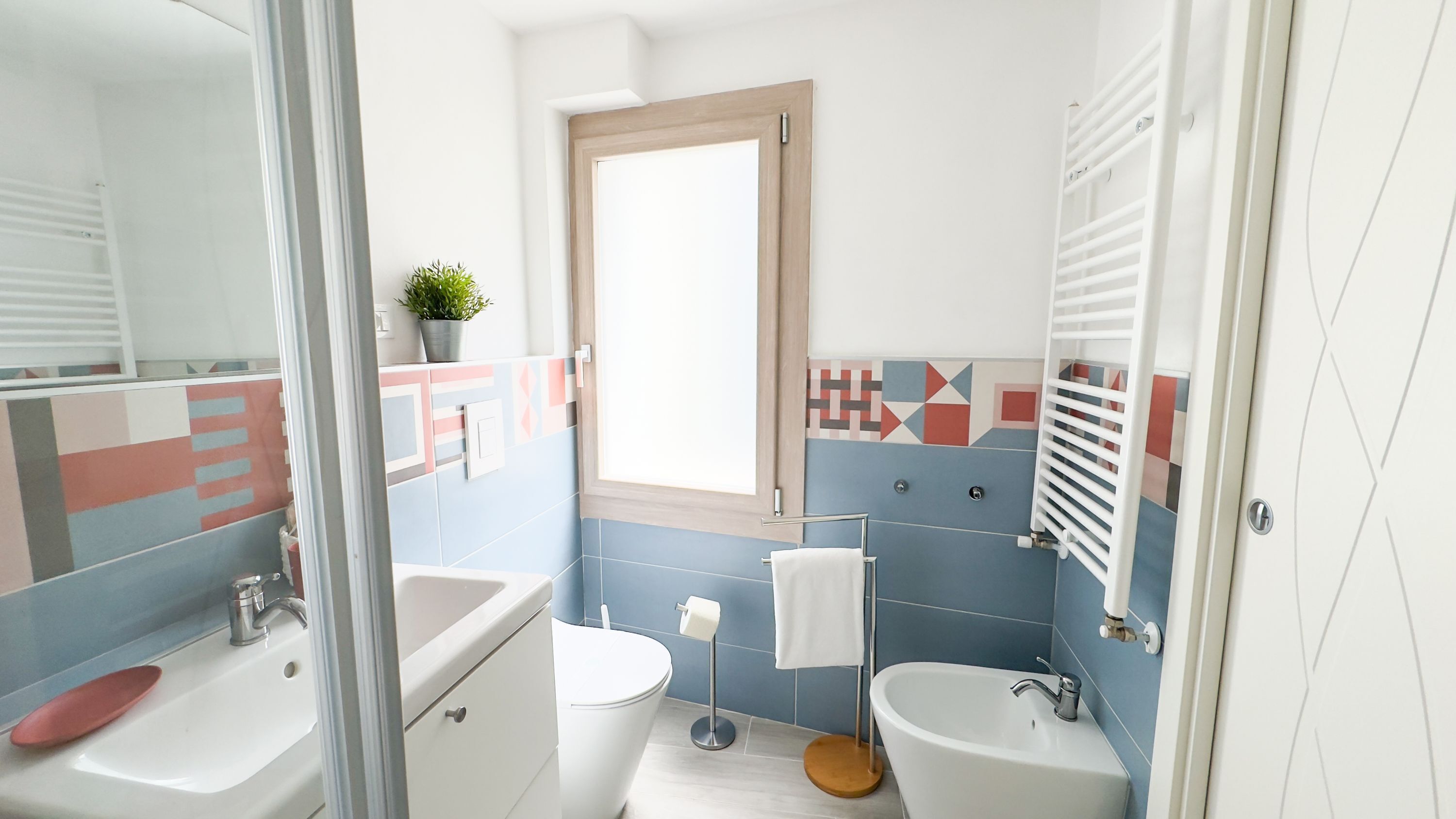 A modern bathroom with a large mirror, two sinks, a toilet, and a window.
