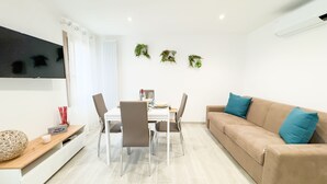 Una sala moderna con un sofá beige, una mesa de comedor con cuatro sillas, un televisor de pantalla plana montado en la pared y tres macetas colgantes.