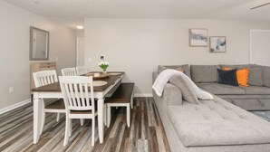 Un área de comedor con una mesa de madera y sillas blancas, un sofá gris con almohadones naranjas y un cuadro en la pared.