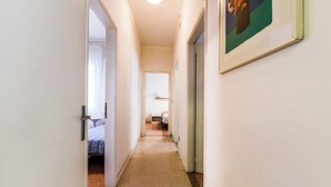 Ein schmaler Flur mit Teppichboden führt zu einem Zimmer mit Bett und Fenster.