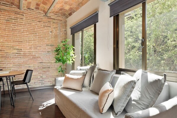 Un salon moderne avec un mur en briques, de grandes fenêtres, un canapé avec des coussins, une table à manger avec des chaises et une plante en pot.