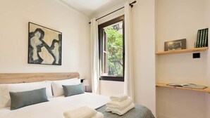 A hotel room with a bed, pillows, a window with curtains, a painting on the wall, and a shelf with books. - La Riera Principal (Barcelona)