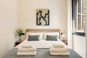 A neatly made bed with white linens and a grey throw, a wall-mounted abstract painting, a bedside table with a lamp, and a window with blinds.