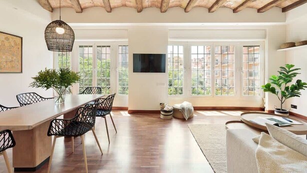 A spacious room with a wooden ceiling, large windows, a dining area with chairs, a sofa, and a TV mounted on the wall. - La Riera Principal (Barcelona)