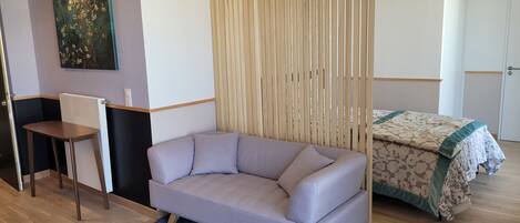A modern hotel room with a grey sofa, a small desk, a wooden partition, and a bed with a patterned bedspread.